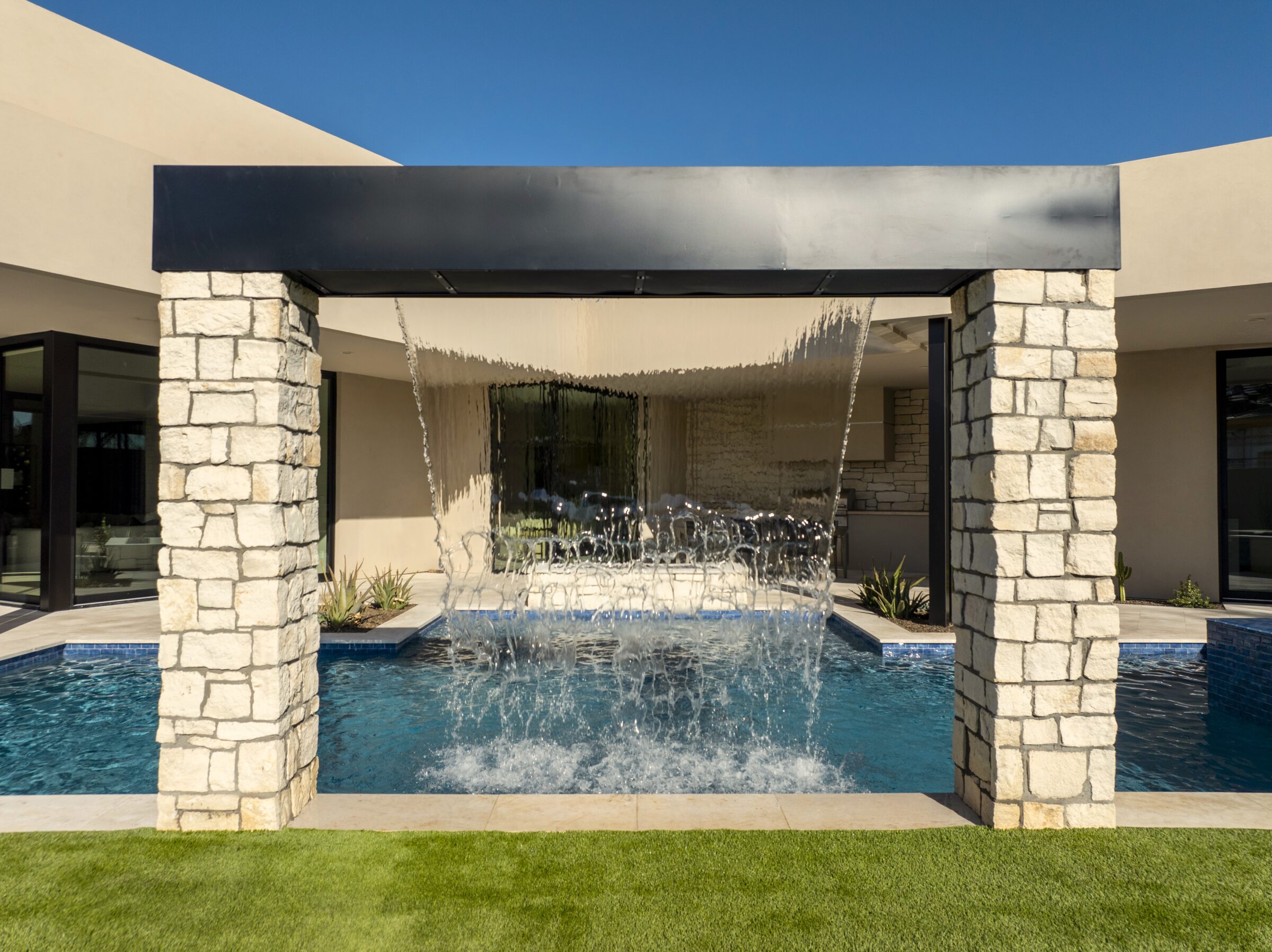 Modern building with a stone archway and waterfall feature over a small pool, surrounded by grass and clear blue sky.