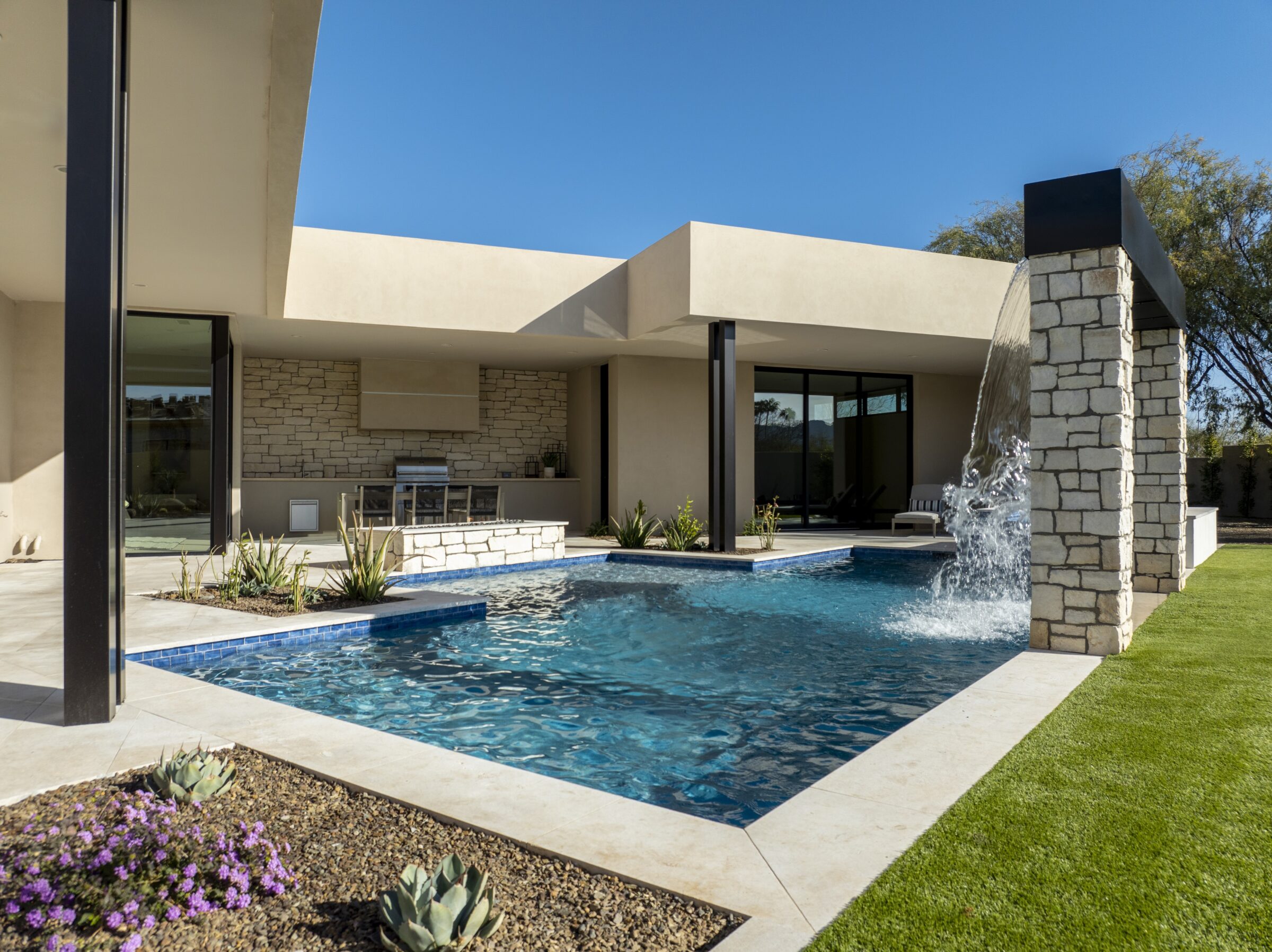 Modern house featuring a rectangular pool, waterfall, and outdoor kitchen. The landscape includes succulents and purple flowers under a clear blue sky.