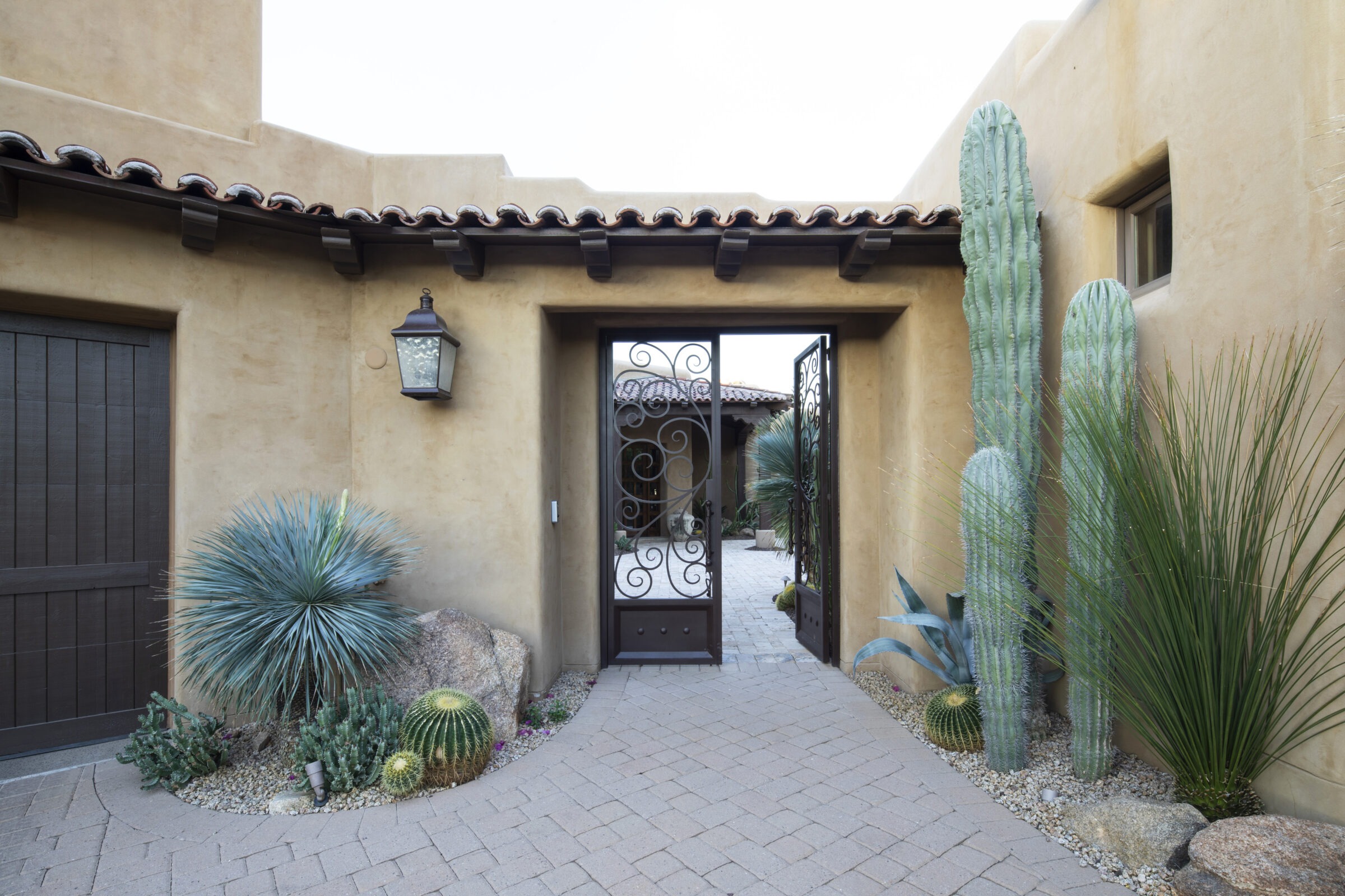 Desert-themed courtyard entrance with stucco walls, wrought iron gate, various cacti, brick paving, and a vintage lantern, creating a Southwestern aesthetic.