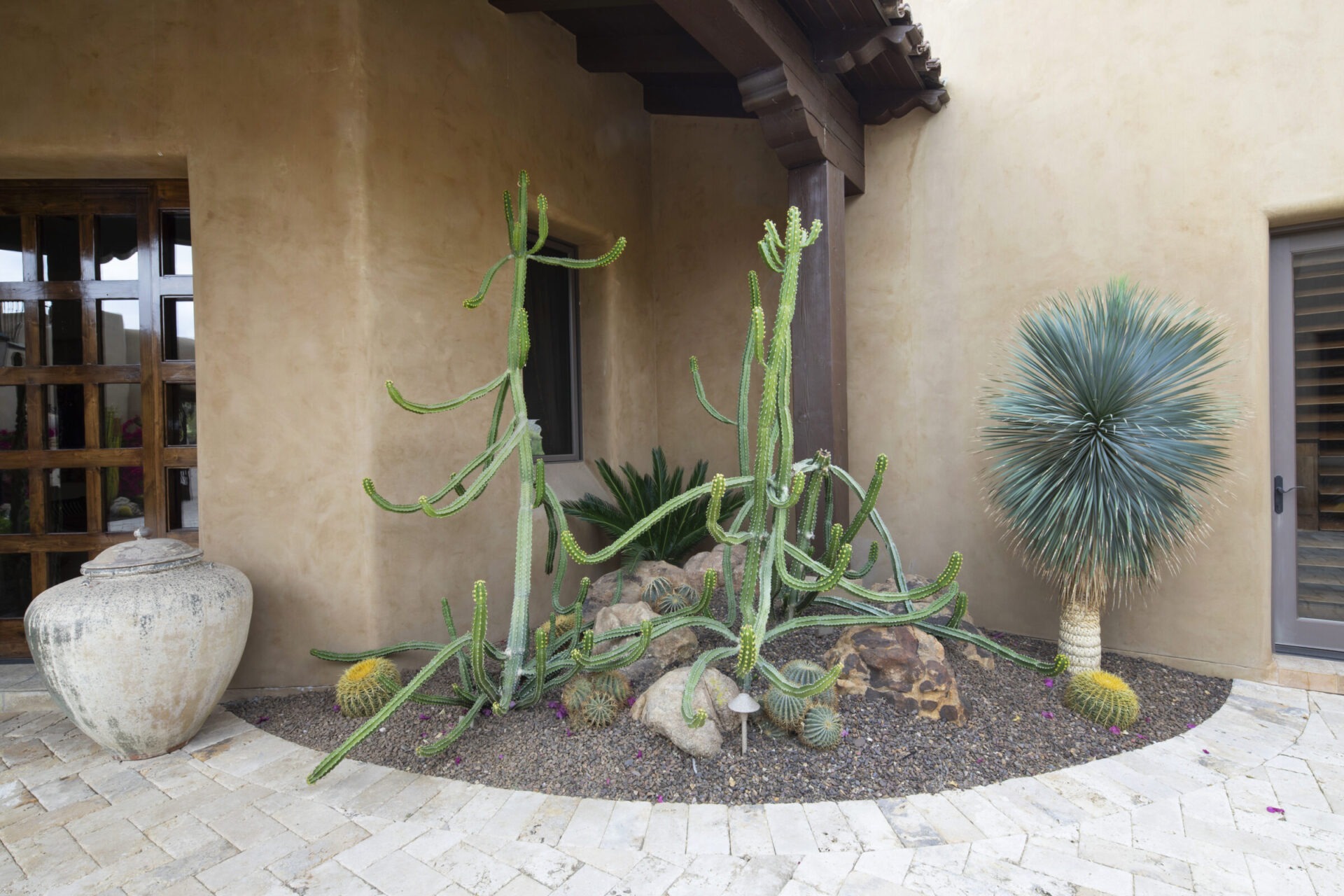 Desert garden with cacti and a yucca plant beside a rustic adobe-style building featuring a large clay pot and wooden beams.