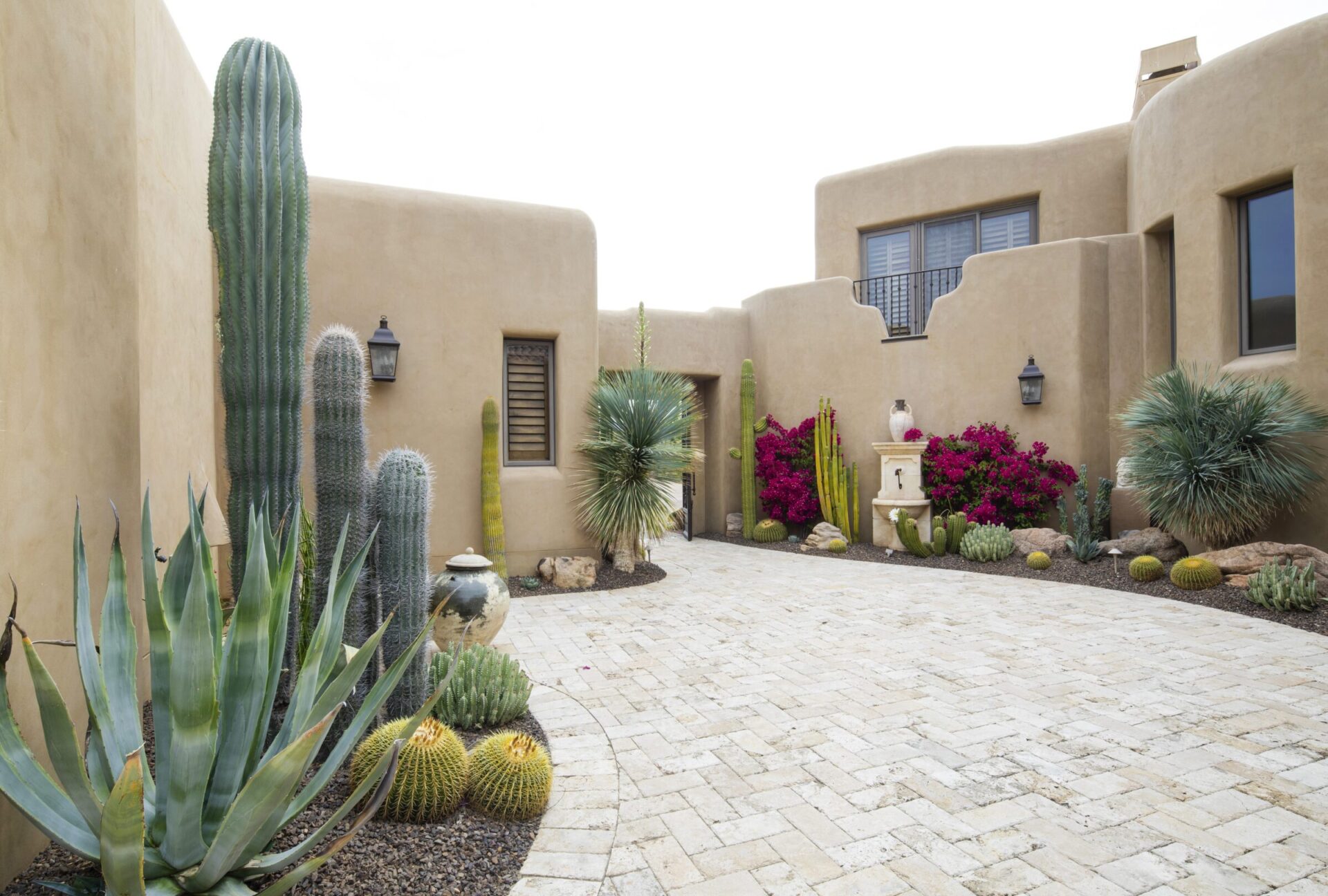 Desert-style courtyard with adobe walls, various cacti, colorful flowers, and traditional pottery. Paved pathway leads through the arid, landscaped garden.