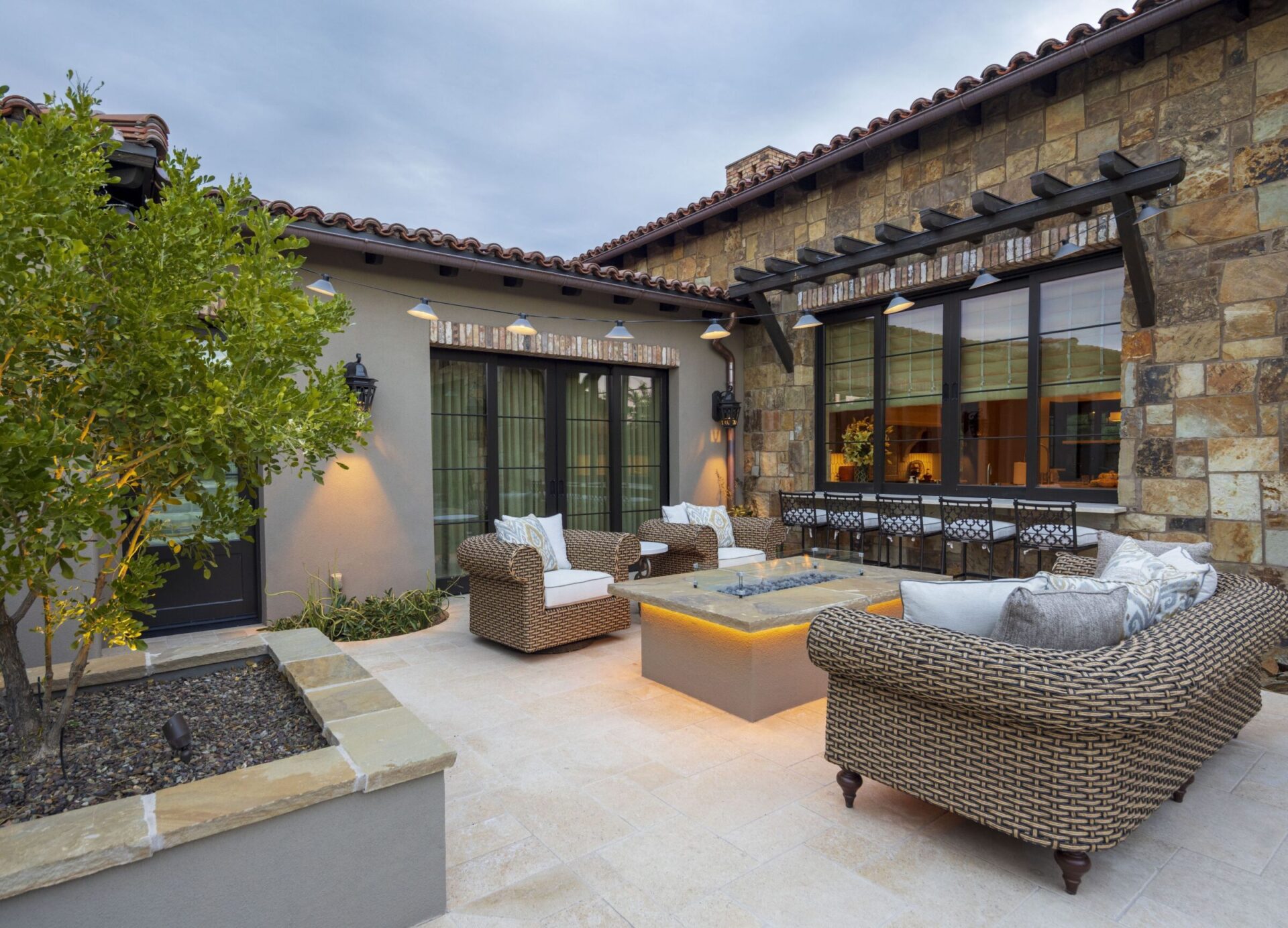 Elegant patio with wicker furniture, stone detailing, and a fire pit. Ambient lighting enhances the cozy outdoor atmosphere next to large windows.