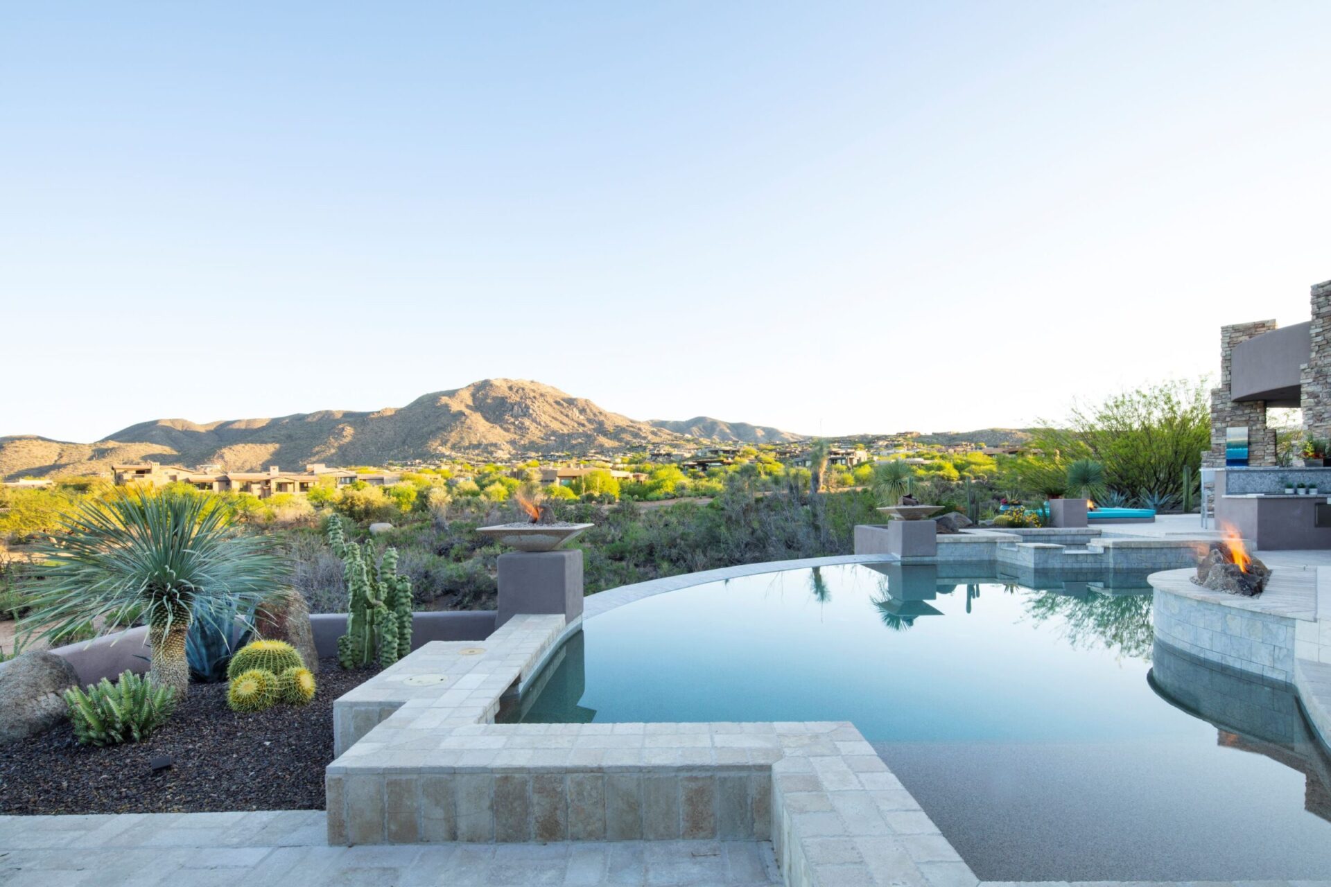 Luxurious infinity pool with fire pit overlooks vast desert landscape, cacti, and distant mountains. Modern architecture and serene setting create a tranquil atmosphere.