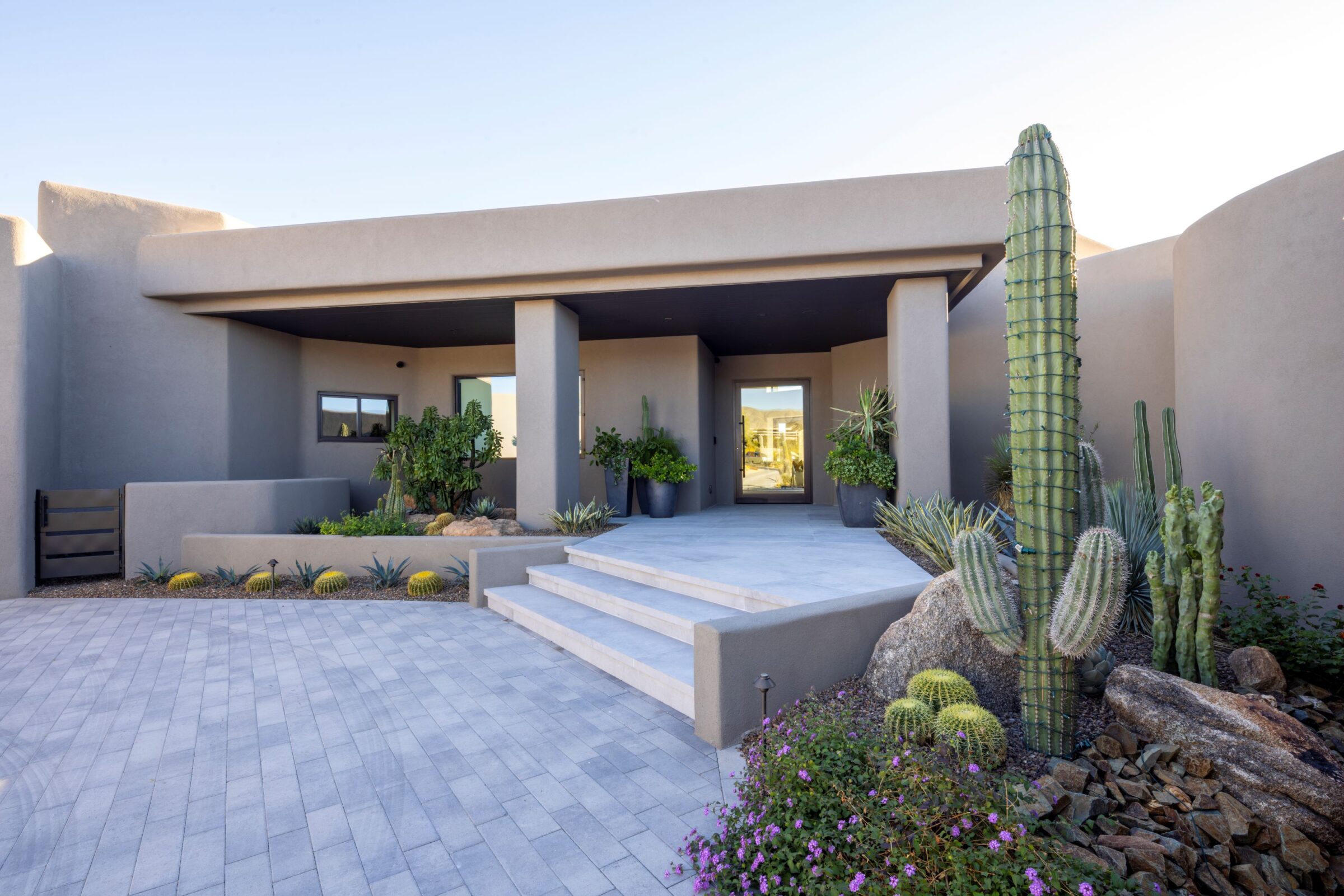 Modern adobe-style house with minimalist design, featuring large cacti and desert landscaping at the entrance under a clear blue sky.