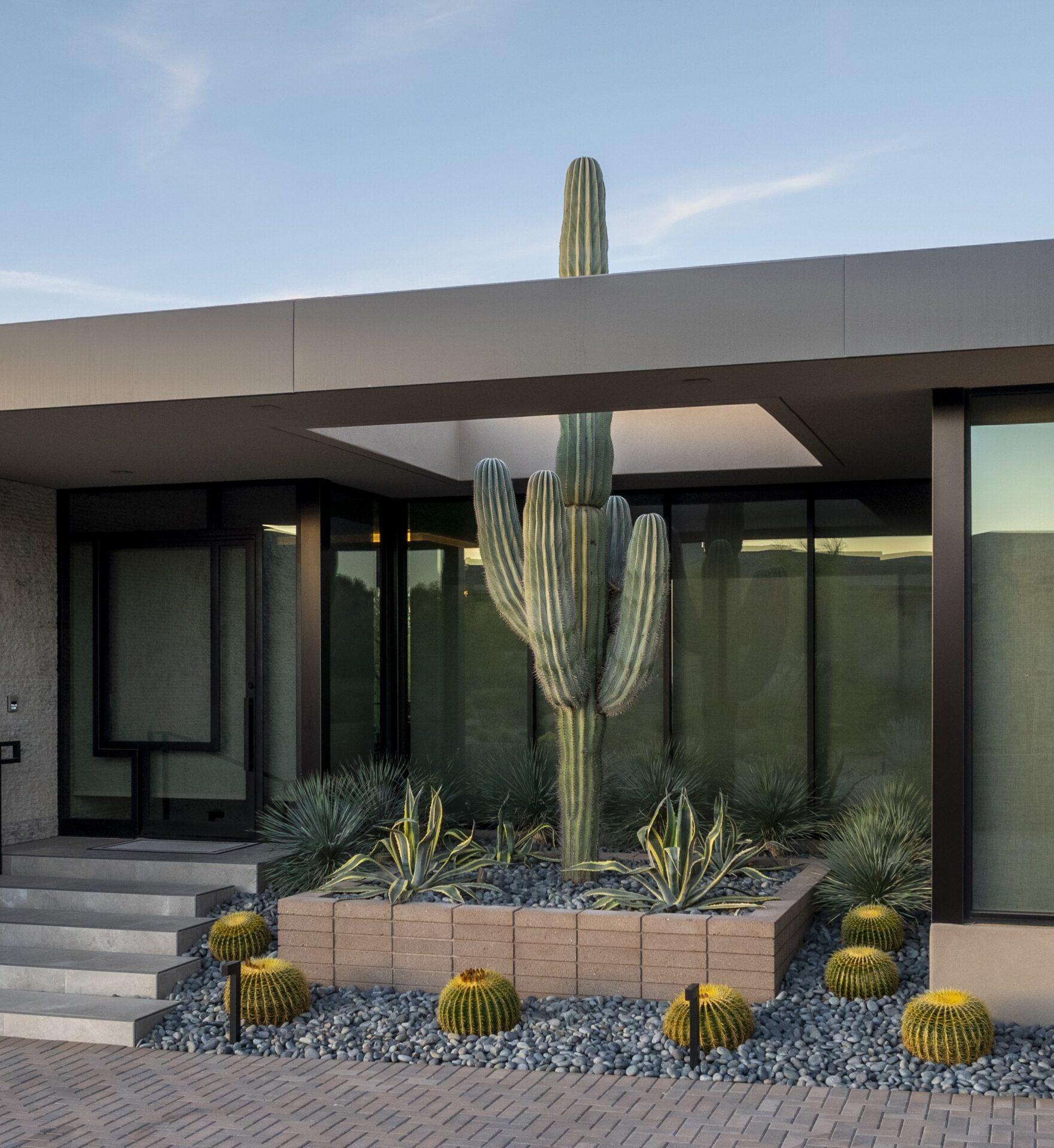 Modern home entrance with large cactus and desert landscaping, featuring neatly arranged stones and succulent plants, under a clear blue sky.