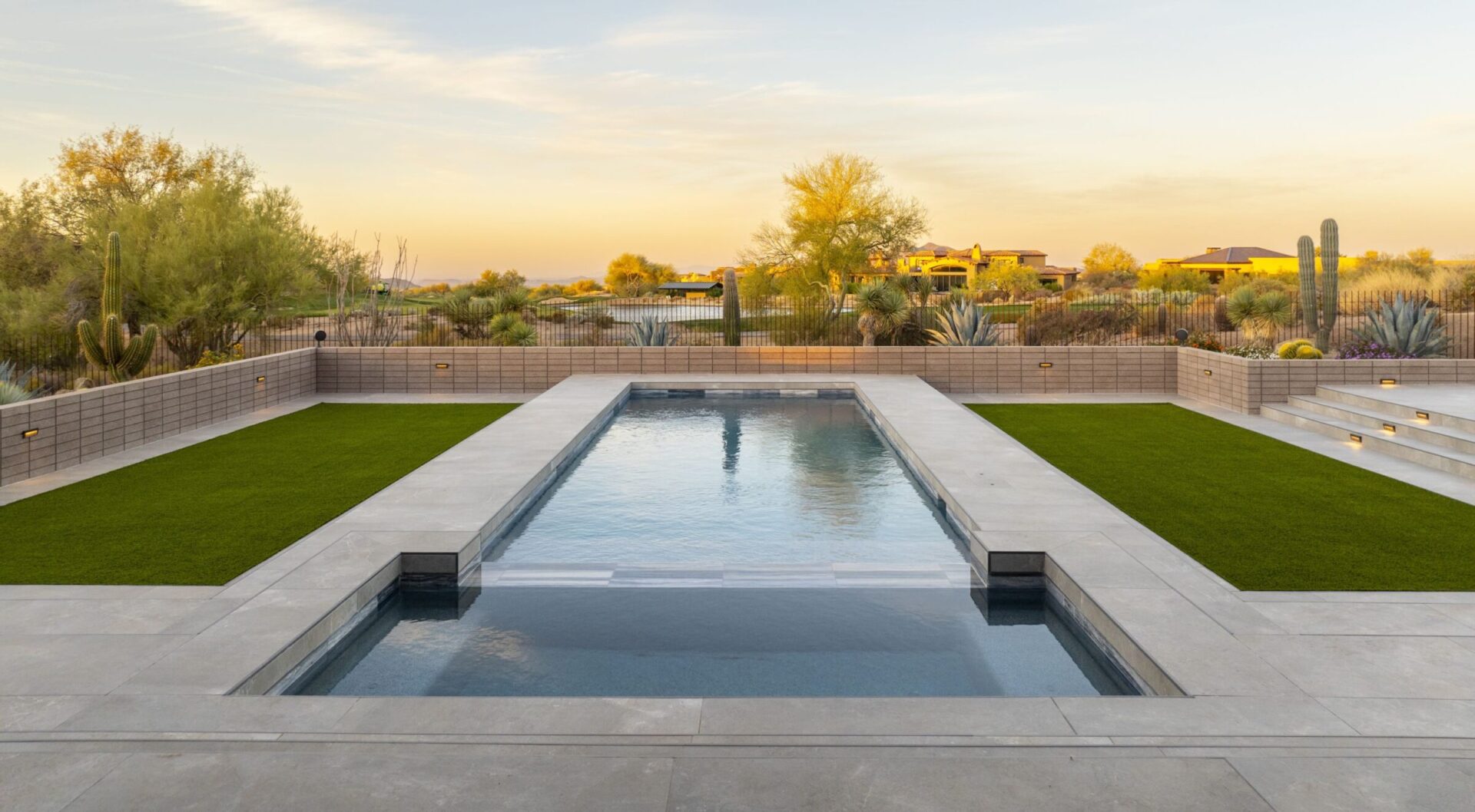 Modern backyard features a rectangular pool surrounded by manicured lawns, cacti, and landscaped greenery under a clear sky at sunset.