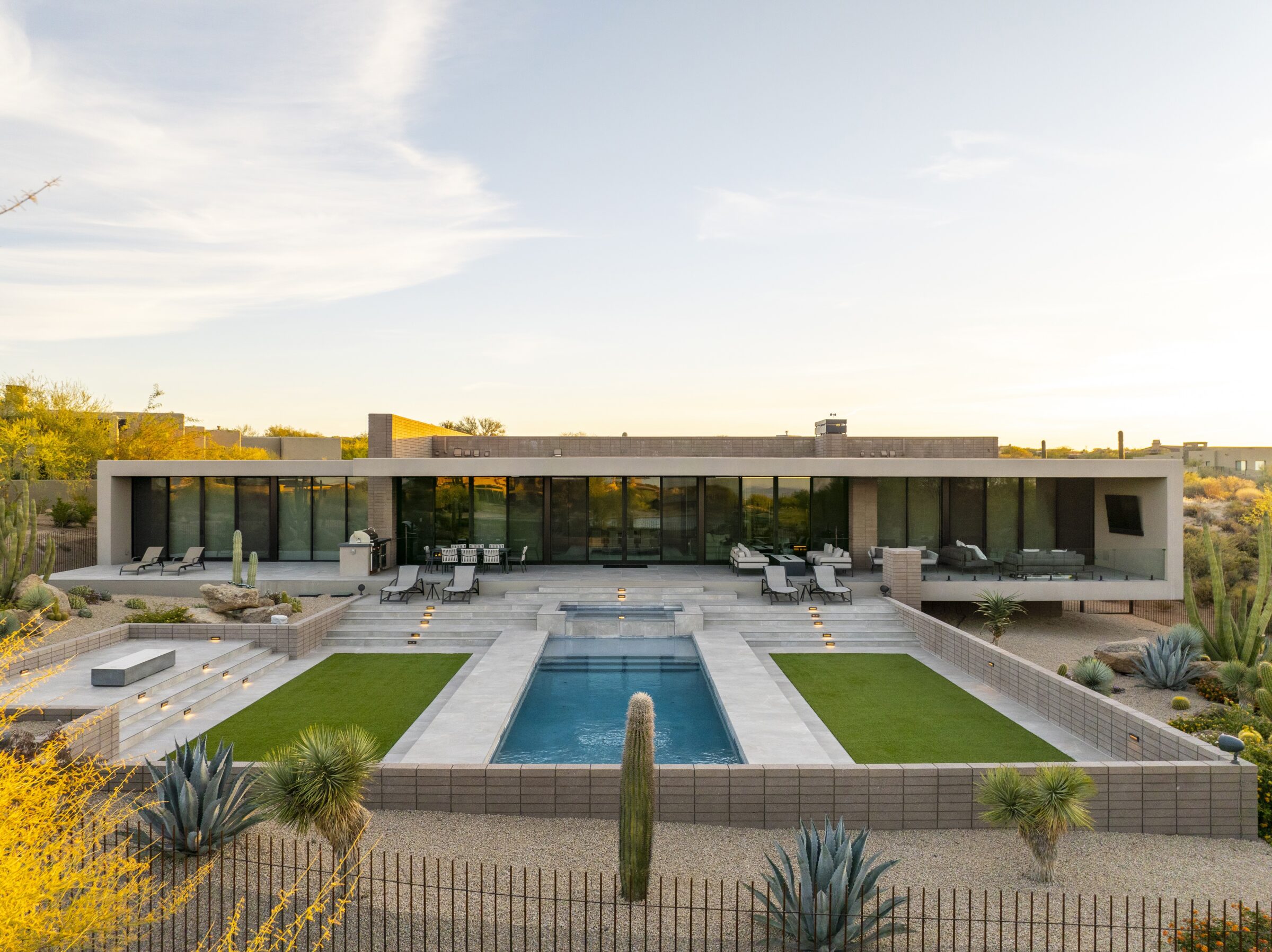 Modern, flat-roofed house with large glass windows, surrounded by desert landscaping. Central pool and patio area overlook a scenic horizon.