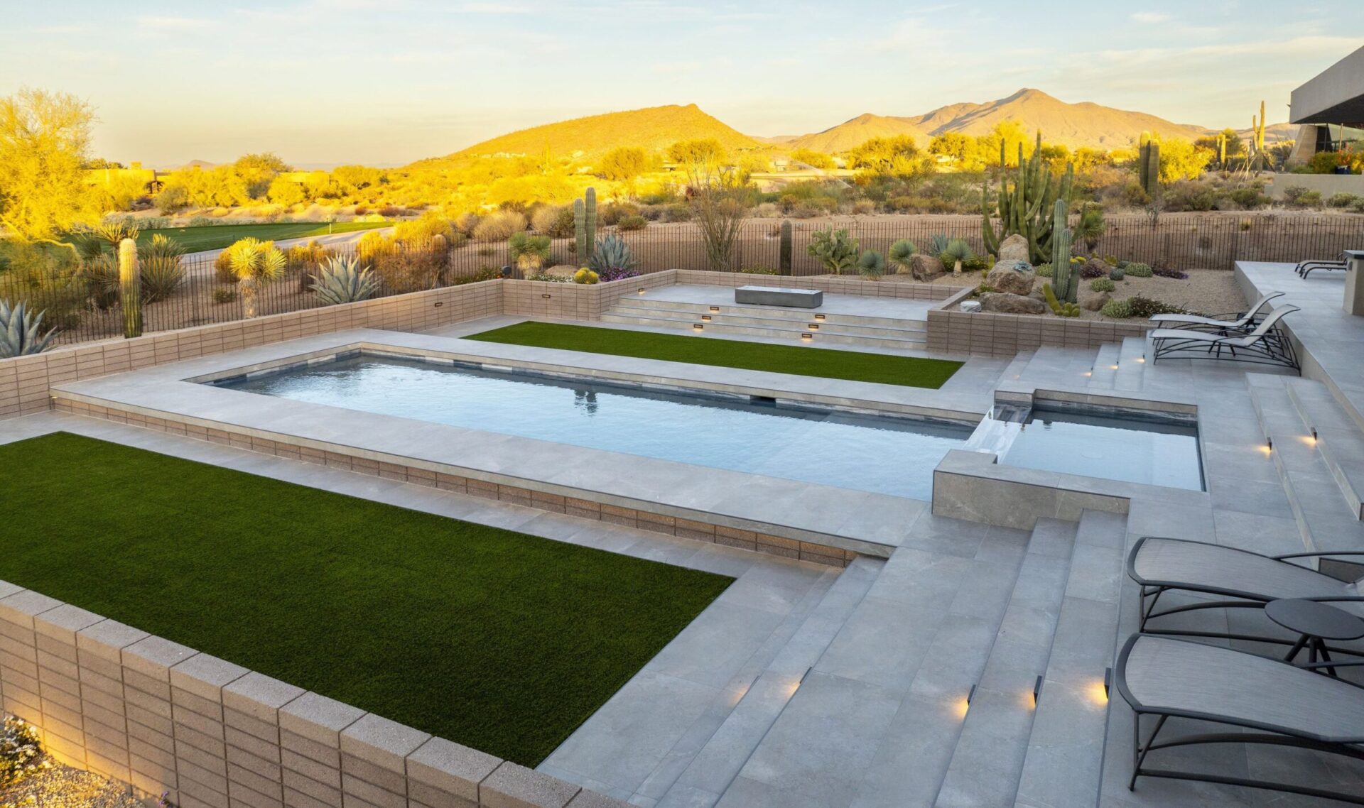 Modern pool with geometric design, surrounded by green lawns and desert landscape. Lounge chairs and steps leading to water. Serene, sunny setting.