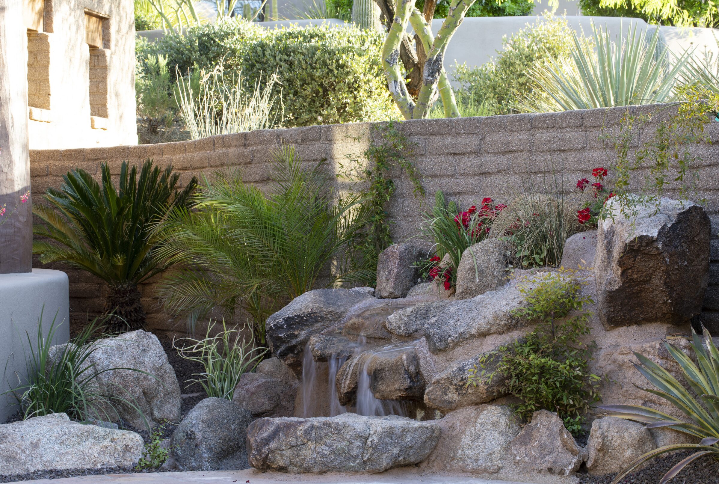 A tranquil garden features a rock waterfall, surrounded by lush plants and flowers, with a stone wall and partial building visible.