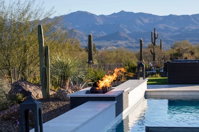 Desert landscape with cacti and a pool featuring a fire pit. Mountains in the background under a clear blue sky.