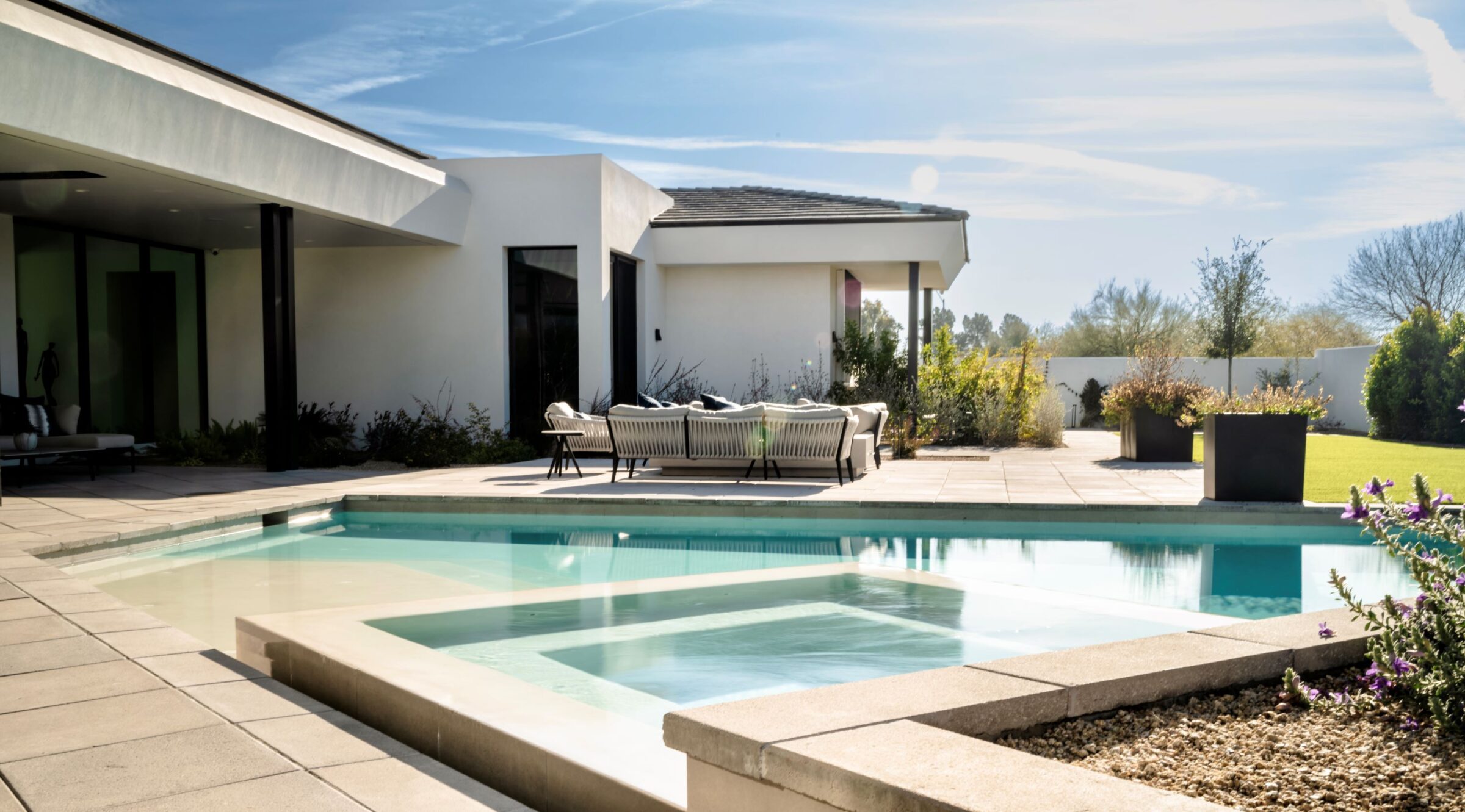 Modern house with a backyard patio, swimming pool, and stylish outdoor furniture surrounded by greenery under a clear, sunny sky.