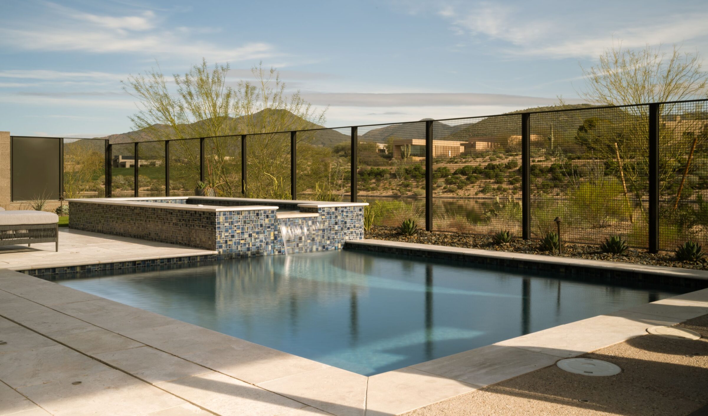 A serene backyard scene featuring a modern swimming pool, tiled waterfall, desert landscape, and mountains under a cloudy sky.