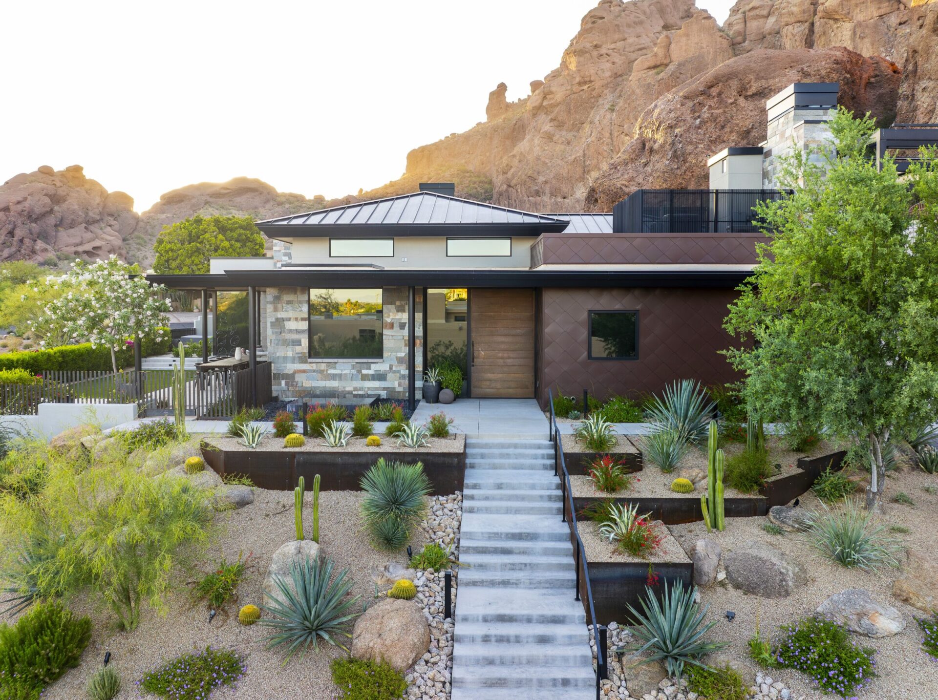 Modern house with stone and metal facade surrounded by desert landscaping, situated near rocky hills under a clear sky.