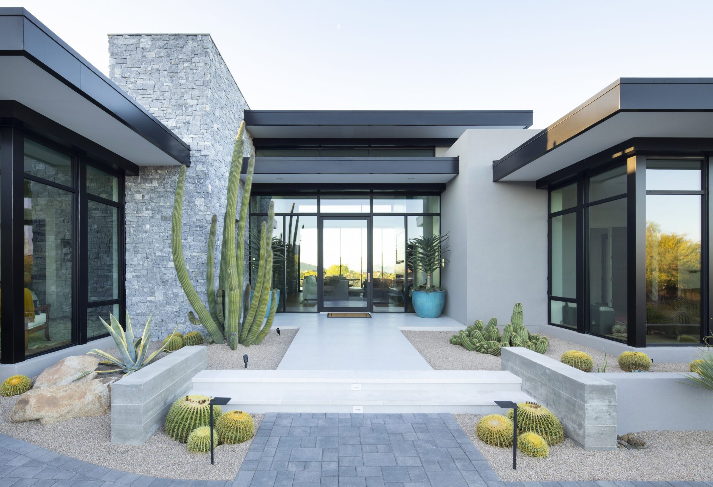 Modern house entrance with large windows, stone walls, and desert landscaping featuring cacti and succulents. Sleek, minimalist architecture with clean lines.