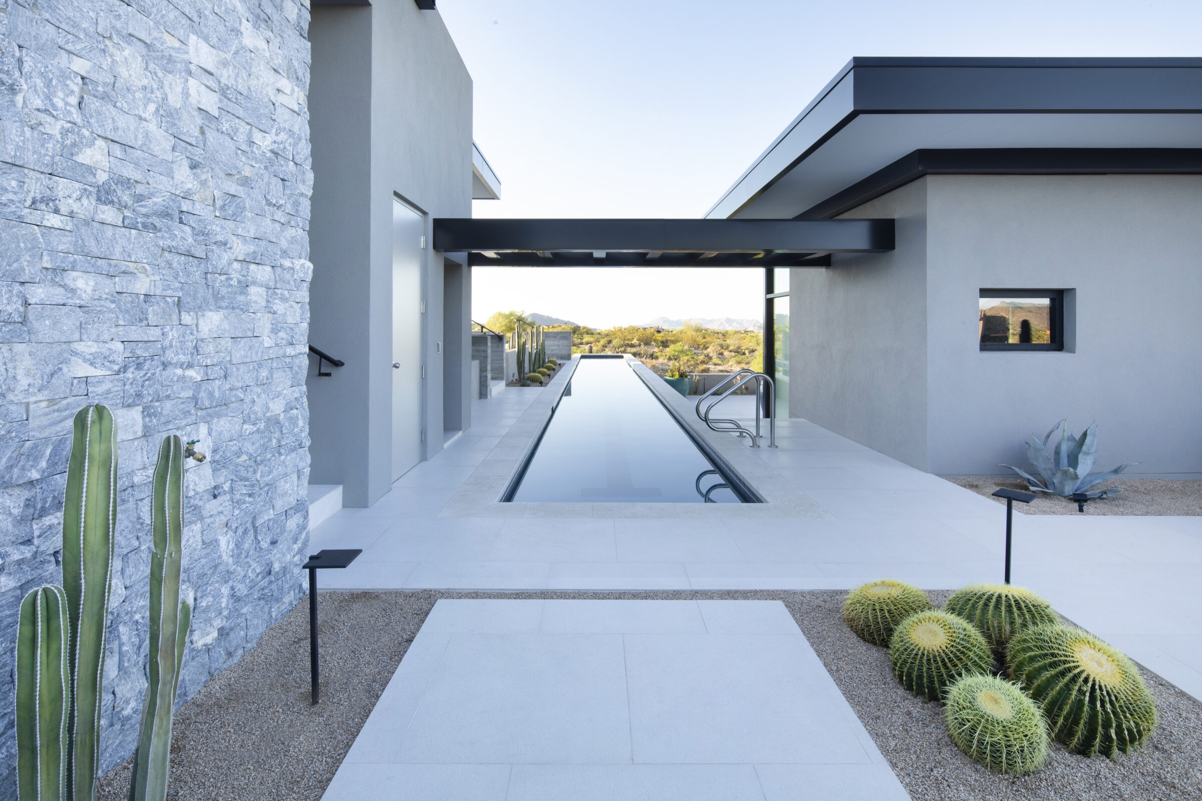 Modern house with sleek design, featuring an elongated pool, stone walls, and desert plants, situated in a scenic landscape with distant mountains.