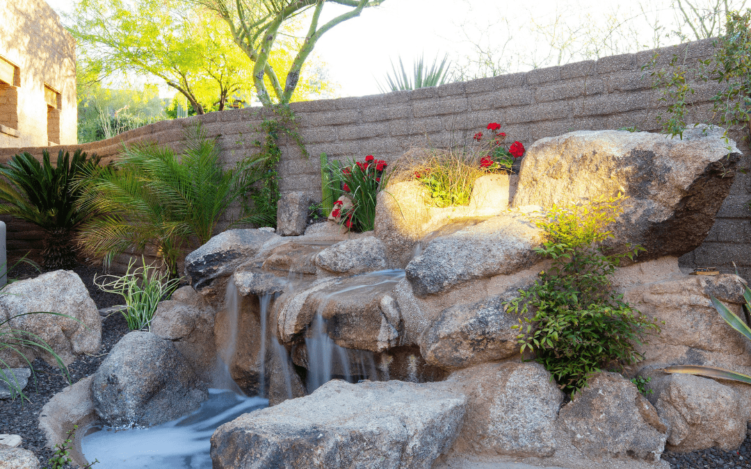 A serene, sunlit backyard features a small waterfall cascading over rocks, surrounded by lush greenery and vibrant flowers against a brick wall.