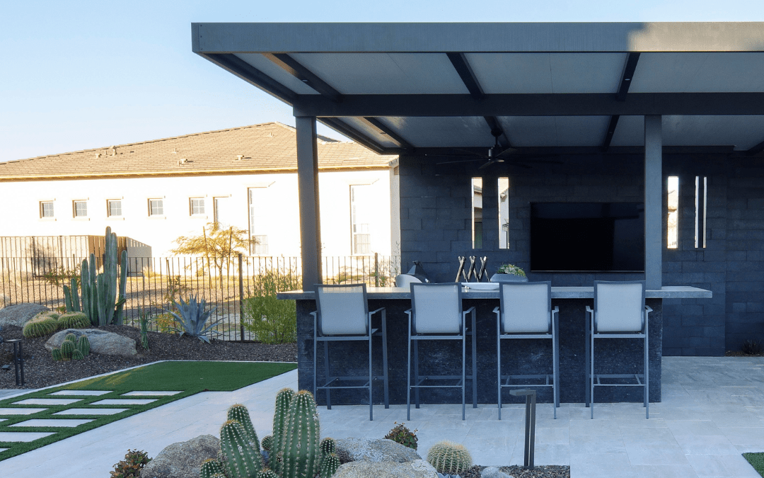 Modern outdoor patio with bar stools, counter, and TV. Surrounded by cacti, rocks, and grass. Nearby building visible in sunlight.