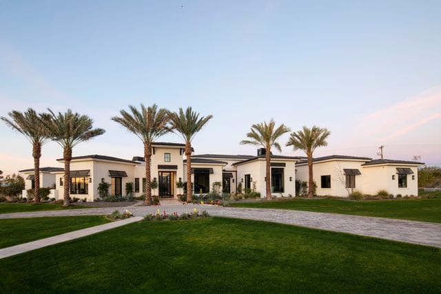 A modern, single-story house with palm trees, expansive lawn, and a stone walkway under a clear sky. No recognizable landmarks or historical buildings.