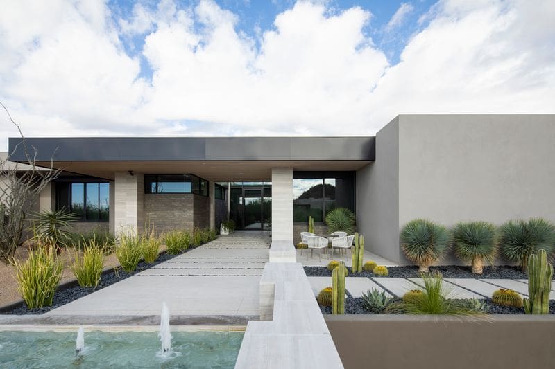 Modern house exterior with minimalist design, desert landscaping, and a water feature. Clean lines and neutral tones under a partly cloudy sky.