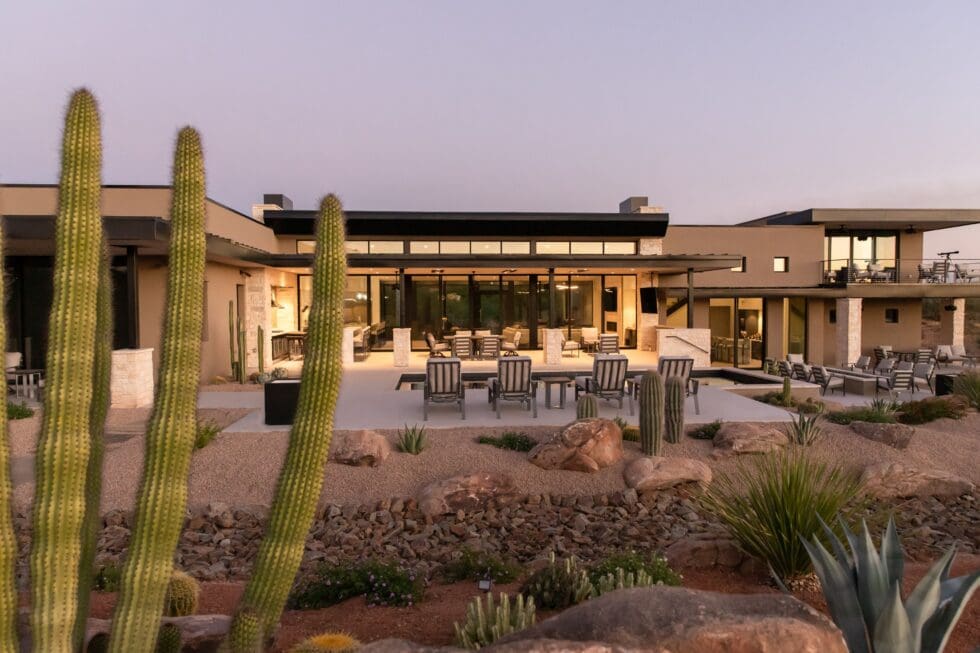 Modern house surrounded by desert landscape, featuring cacti and contemporary outdoor seating under a clear sky, highlighting minimalistic architectural design.