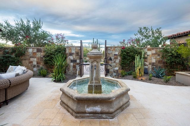 Stone fountain in a garden courtyard with desert plants and tiled walls, under a cloudy sky. Outdoor seating is visible on the left.