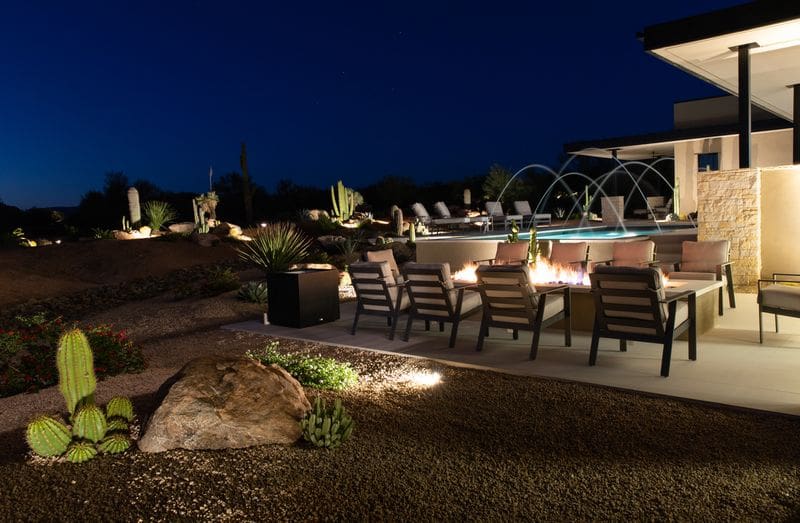 Modern outdoor patio with fire pit, stylish seating, and desert landscaping. Nighttime setting with cacti under a starry sky, near a contemporary pool.