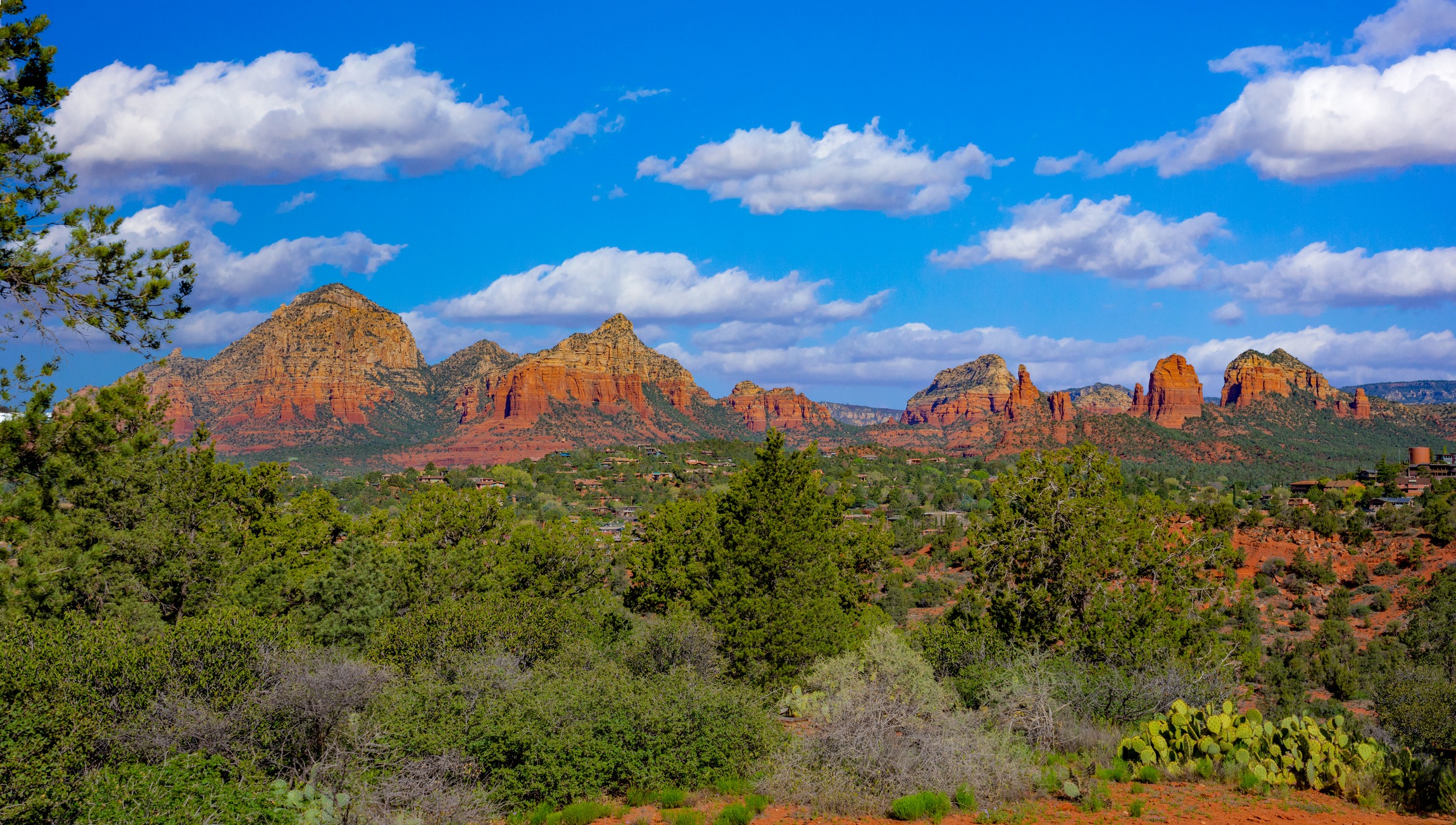 A scenic view of Sedona's red rock formations under a bright blue sky with scattered clouds, surrounded by lush greenery and desert plants.