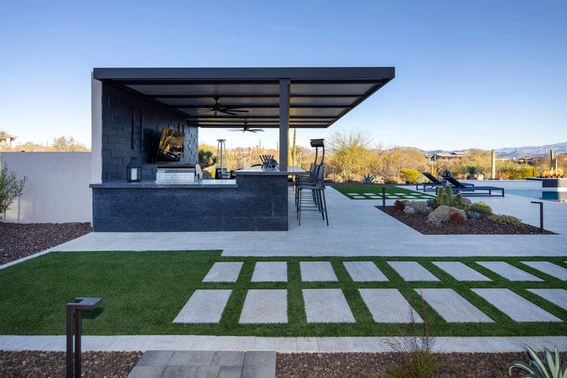 Modern outdoor kitchen with bar stools, TV, and ceiling fans. Surrounded by landscaped garden, paving stones, and desert scenery under a clear blue sky.