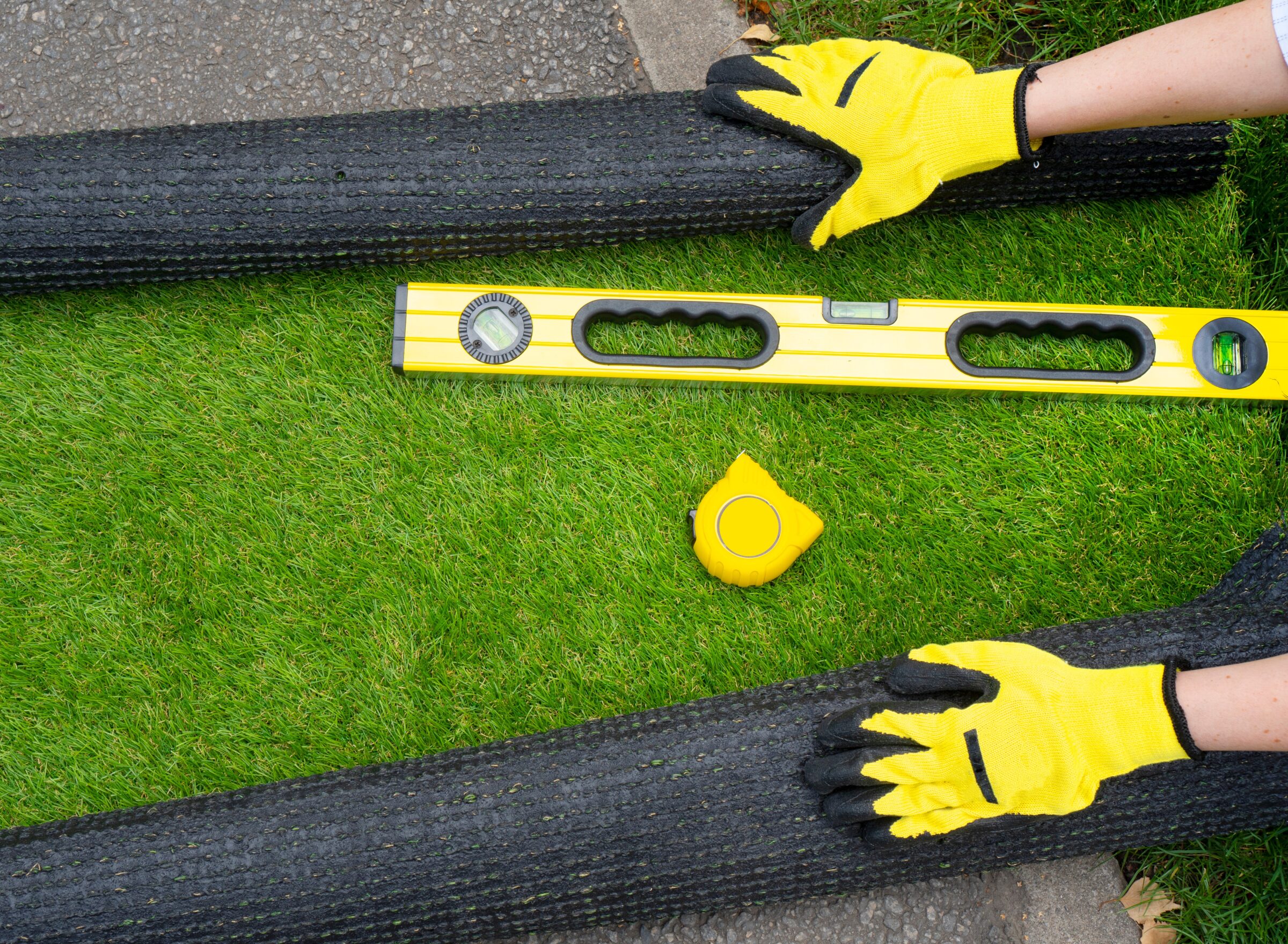 Person installing artificial grass with yellow gloves, using a spirit level and tape measure on a concrete surface.