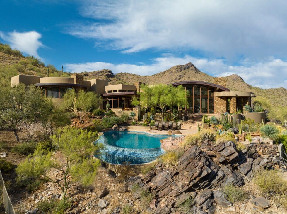 Modern desert home with large windows, surrounded by rocky terrain and greenery. Features an infinity pool, blending with the natural landscape.